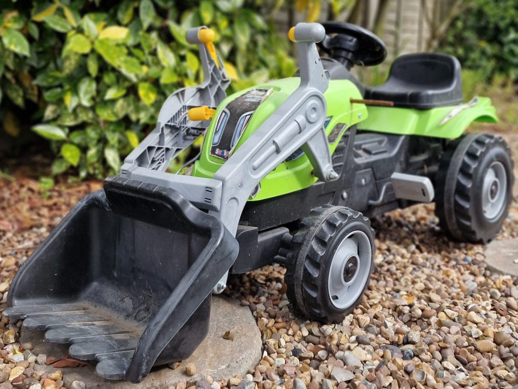A colorful green toy tractor with a front loader parked on a gravel surface, surrounded by greenery.