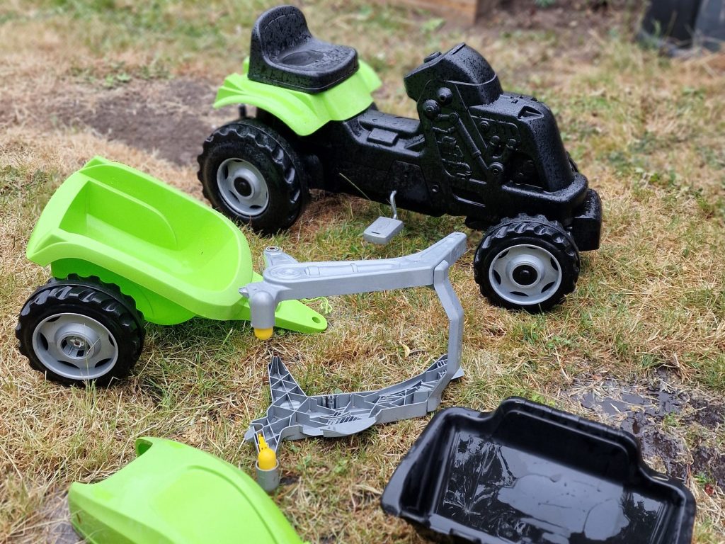 A green toy truck and tipper trailer on grass, with additional toy parts scattered nearby.