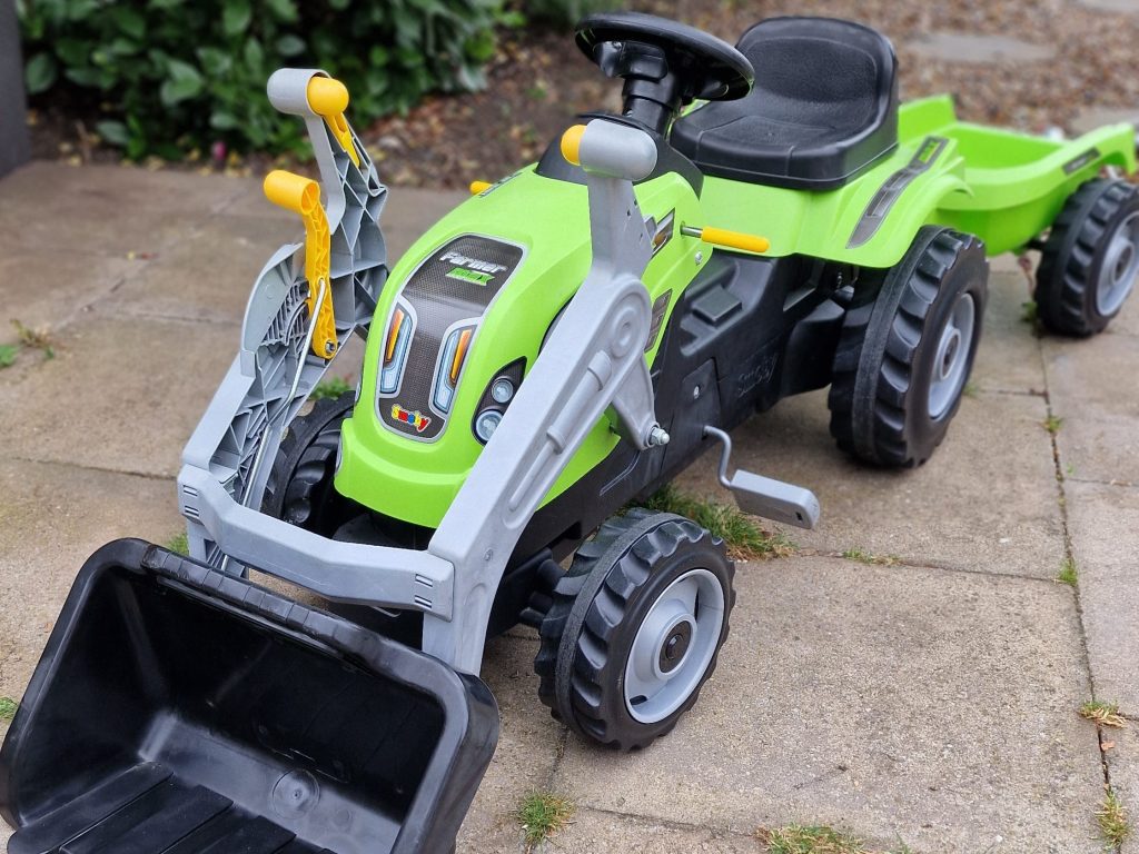 A green toy tractor with a black bucket, parked on a paved surface surrounded by greenery.